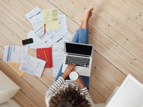 View from above of unrecognizable Afro woman reads information from paper documents, checks database on modern laptop computer, works freelance in modern apartment on floor, drinks hot coffee